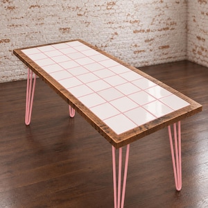 Peut inclure: Une table rectangulaire dotée d'un plateau composé d'une grille de carreaux carrés rose clair, encadrée par une bordure en bois brun foncé. La table est soutenue par quatre pieds en épingle fins et en forme de U, de couleur rose clair mat.