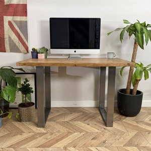 May include: A wooden desk with metal legs. The desk has a light wood finish and the legs are a silver colour. There is a computer monitor, a potted plant, and a cup on the desk. The desk is sitting on a wood floor with a herringbone pattern.