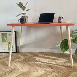 Pode incluir: Uma pequena mesa laranja com pernas de madeira clara. A mesa tem um laptop, uma caneca, uma planta e um pote em cima. A mesa está sobre um piso de madeira com um padrão de espinha de peixe.