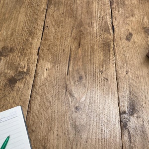K&ouml;nnte beinhalten: Nahaufnahme einer Holzoberfl&auml;che mit einer hellbraunen Beize. Die Oberfl&auml;che hat eine nat&uuml;rliche Holzmaserung und besteht aus mehreren Holzbrettern. Eine kleine Topfpflanze mit einer Sukkulente befindet sich in der oberen rechten Ecke des Bildes. Ein Notizbuch mit einem gr&uuml;nen Stift befindet sich in der unteren linken Ecke des Bildes.