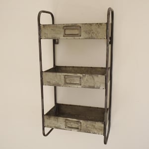 May include: A three-tiered, wall-mounted storage shelf with a distressed, galvanized metal finish. The shelf has a dark metal frame with curved top supports and rectangular label holders on each shelf. The shelves are approximately 12 inches wide.