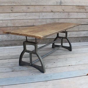 Op de afbeelding: Een rustieke houten tafel met een metalen onderstel. De tafel heeft een rechthoekig blad en een zwart metalen frame met twee dwarsbalken. De tafel staat op een houten vloer.