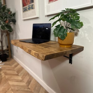 K&ouml;nnte beinhalten: Ein Wandregal aus Holz mit einer lebenden Pflanze in einem gelben Topf und einem Laptop darauf. Das Regal ist aus Naturholz mit einer lebenden Kante und an einer wei&szlig;en Wand befestigt. Das Regal wird von einer schwarzen Metallhalterung getragen.