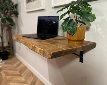 May include: A wooden wall-mounted shelf with a live plant in a yellow pot and a laptop computer on top. The shelf is made of natural wood with a live edge and is mounted to a white wall. The shelf is supported by a black metal bracket.