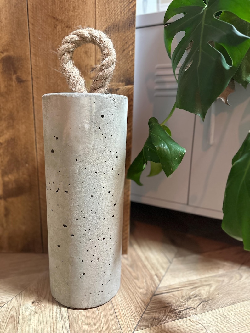 May include: A grey concrete doorstop with a rope handle. The doorstop is sitting on a wooden floor.
