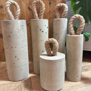 May include: Five concrete doorstops with rope handles. The doorstops are of varying sizes and are arranged in a row on a wooden floor.