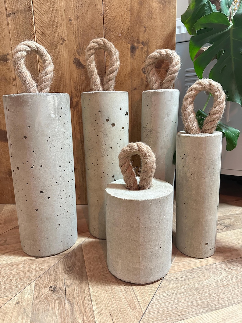 May include: Five concrete doorstops with rope handles. The doorstops are of varying sizes and are arranged in a row on a wooden floor.