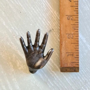 Puede incluir: Un tope de puerta de metal con forma de mano de color gris oscuro. La mano está levantada con los cinco dedos extendidos.