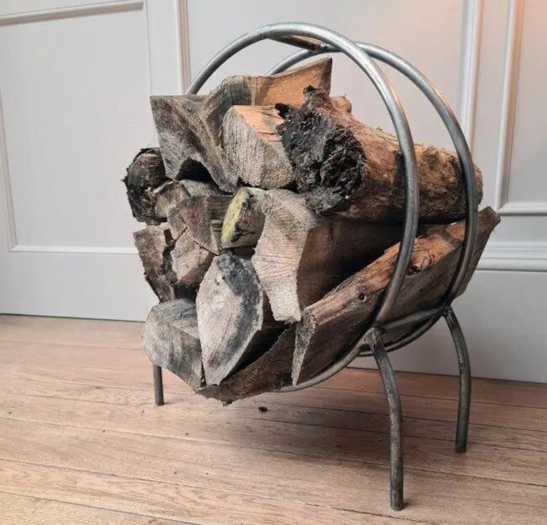 Puede incluir: Un porta le&ntilde;a de metal plateado con un dise&ntilde;o circular y tres patas. El porta le&ntilde;a est&aacute; lleno de una pila de le&ntilde;a.