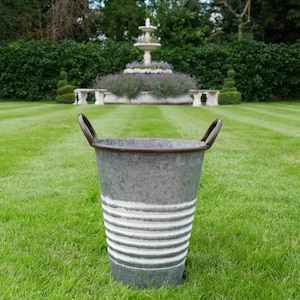 Puede incluir: Un cubo de metal galvanizado gris con dos asas arqueadas. El cubo tiene rayas blancas horizontales y un borde oscuro. Está sobre un césped verde, con una fuente y setos en el fondo.