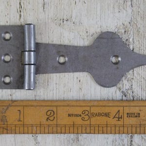 Könnte beinhalten: Ein graues Metallscharnier mit dekorativem spitzem Ende, mit einer rechteckigen Montageplatte mit drei Löchern und einem Lineal. Das Scharnier ist etwa 13 cm lang.