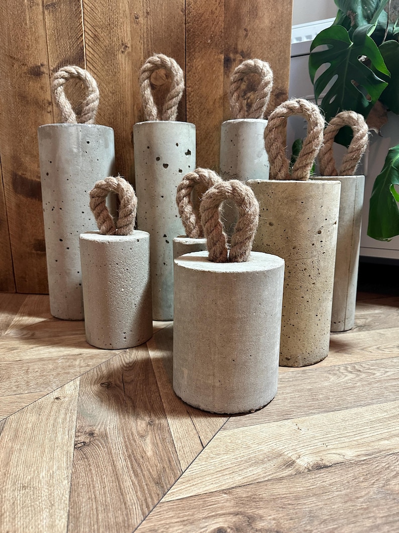 May include: A set of six concrete doorstops with rope handles. The doorstops are various sizes and are arranged on a wooden floor.