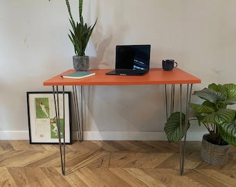 May include: A bright orange desk with hairpin legs. A laptop computer and a mug are on the desk. A potted plant is on the left side of the desk and another potted plant is on the right side of the desk.