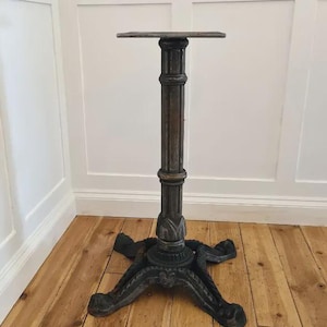 May include: A dark grey cast iron table base with an ornate design. The base has a central column and a four-footed base. The top is a flat, square surface. Suitable for a small table. The base is approximately 76 cm tall.