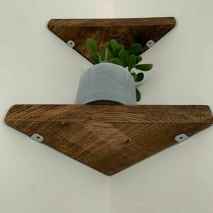 May include: Two wooden triangular shelves mounted in a corner, with a small potted plant on the top shelf. The shelves are stained a dark brown colour and have metal brackets.