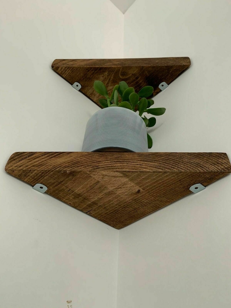 May include: Two wooden triangular shelves mounted in a corner, with a small potted plant on the top shelf. The shelves are stained a dark brown colour and have metal brackets.