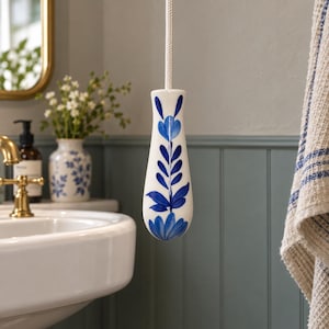 May include: A white ceramic light pull with a tapered, rounded shape featuring a hand-painted blue floral pattern. The pull is attached to a thick, textured white cord that extends upwards, providing a functional handle for operating light switches.