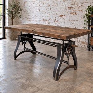 May include: A rectangular table with a thick, weathered brown wooden top and a black metal base. The base features an adjustable mechanism with a hand crank on the side, connected to a central support bar and curved legs with a distressed finish.