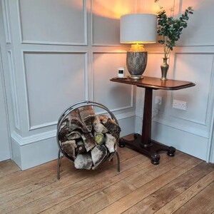 Puede incluir: Un porta-le&ntilde;a de metal con una manta de patr&oacute;n marr&oacute;n y blanco en su interior. El soporte est&aacute; en una habitaci&oacute;n con paredes blancas en panel y suelo de madera. Una mesa peque&ntilde;a con una l&aacute;mpara y un jarr&oacute;n de vegetaci&oacute;n est&aacute; en el fondo.