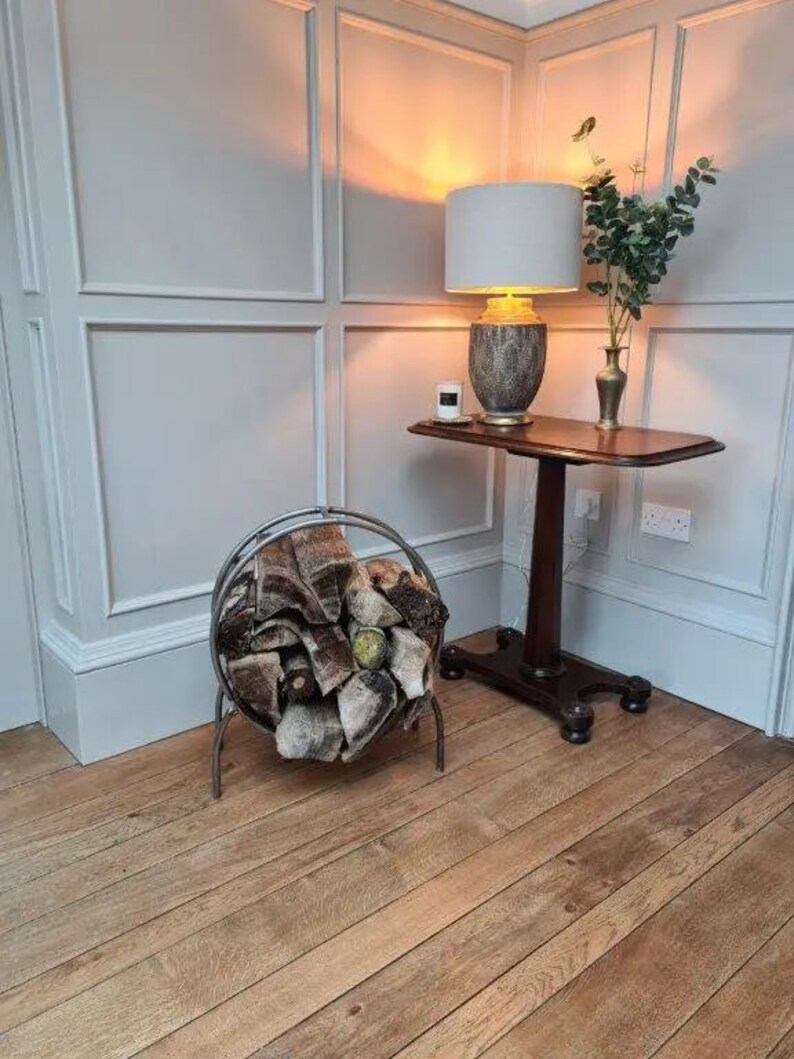 Puede incluir: Un porta-le&ntilde;a de metal con una manta de patr&oacute;n marr&oacute;n y blanco en su interior. El soporte est&aacute; en una habitaci&oacute;n con paredes blancas en panel y suelo de madera. Una mesa peque&ntilde;a con una l&aacute;mpara y un jarr&oacute;n de vegetaci&oacute;n est&aacute; en el fondo.