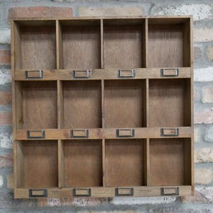 May include: A wooden wall-mounted organiser with multiple compartments. The organiser is made of light brown wood and features small metal label holders on each compartment. The background is a brick wall.