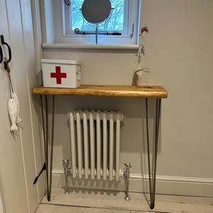 Op de afbeelding: Een rustieke houten consoletafel met haarspeldpoten. De tafel is versierd met een rode EHBO-doos, een ronde spiegel en een decoratieve metalen haan.