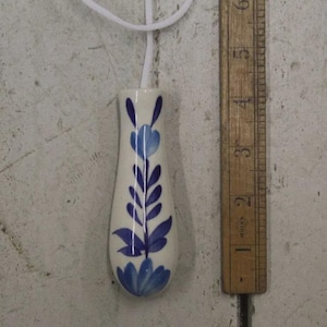 Op de afbeelding: Een witte keramische lichtschakelaar met een blauw bloemmotief. De trekker is langwerpig met een afgeronde bodem en is verbonden met een wit koord. Een houten liniaal ligt ernaast.