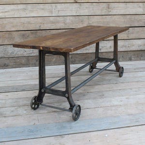 May include: A rectangular table featuring a dark brown wooden top with a visible grain pattern. The base is constructed from dark grey metal, consisting of four vertical supports connected by horizontal bars, each mounted on small circular wheels.