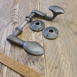 May include: A set of antique-style door hardware in a weathered gray finish. The set includes two door handles, two circular backplates, and a connecting rod. A wooden ruler is in the foreground. The hardware is made of metal.