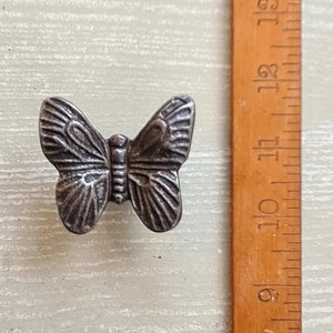 May include: A small, silver-coloured butterfly-shaped decorative knob. The knob is made of metal and has detailed wing and body features. A wooden ruler is next to the knob, showing measurements in inches.