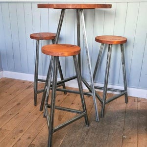 Può includere: Un tavolo da bar rustico con tre sgabelli abbinati. Il tavolo ha un piano rotondo in legno e gambe in metallo. Gli sgabelli hanno sedili rotondi in legno e gambe in metallo. Le gambe in metallo hanno una finitura argentata.