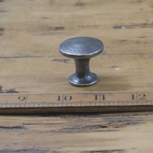 May include: A small, antique-style cabinet knob with a dark, metallic finish. The knob has a rounded top and a short, cylindrical base. It is set against a wooden surface with measurement markings.