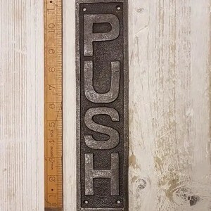 May include: A silver metal sign with the word "PUSH" embossed in raised letters. The sign is mounted on a white wooden surface.