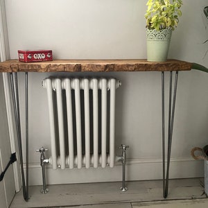 Op de afbeelding: Een rustieke houten consoletafel met een live edge-blad en haarspeldpoten. De tafel is versierd met een rode doos met het woord "OXO" erop. Achter de tafel staat een witte radiator.