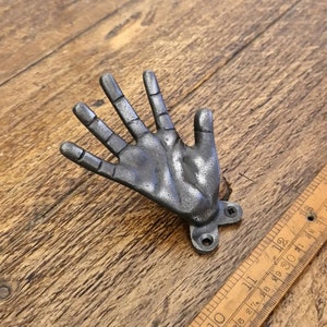 May include: A silver metal hand-shaped hook with a textured surface. The hook is attached to a wooden surface with two screws.