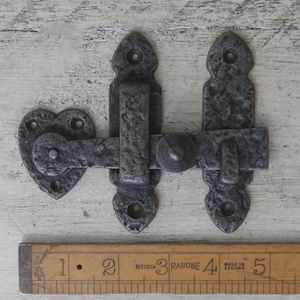 May include: A rustic, dark gray metal latch with a heart-shaped catch and decorative, pointed ends. The latch is shown with a wooden ruler, with the text "RABONE" and "MADE IN ENGLAND" visible.