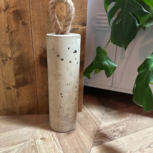 May include: A concrete doorstop with a rope handle. The doorstop is cylindrical in shape and has a rough, textured surface.