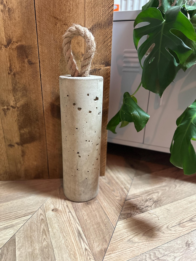 May include: A concrete doorstop with a rope handle. The doorstop is cylindrical in shape and has a rough, textured surface.
