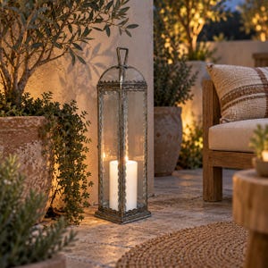 May include: A tall, rectangular metal lantern with clear glass panels and a decorative scalloped trim along the edges. The lantern features a top handle and contains a single white pillar candle that is lit, emitting a warm, soft glow.