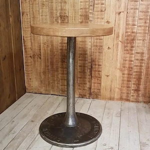 May include: A round wooden table top with a cast iron base. The base is a large, round disc with a raised centre post and a decorative design around the edge. The table is in a room with wood panelling on the walls.