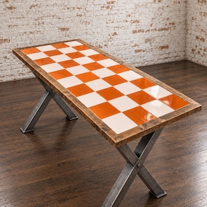 May include: A rectangular table featuring a tabletop with a glossy orange and white chequered tile pattern framed by a rustic brown wooden border. The table is supported by two intersecting X-shaped metal legs with a dark grey industrial finish.