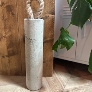 May include: A grey concrete doorstop with a rope handle. The doorstop is sitting on a wooden floor.