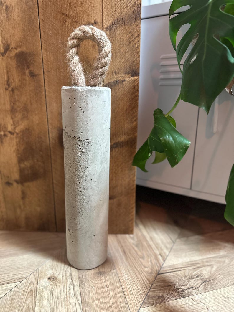 May include: A grey concrete doorstop with a rope handle. The doorstop is sitting on a wooden floor.