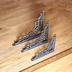 May include: Three decorative, Art Deco-style metal shelf brackets in varying sizes. The brackets are dark gray with geometric cut-out designs. They are arranged on a wooden surface with a ruler.