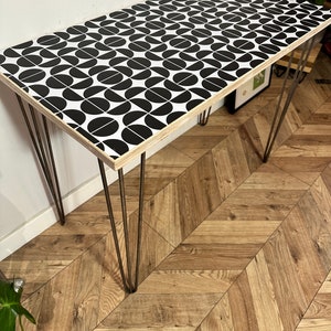 Puede incluir: Una mesa con una tapa de madera con un patrón geométrico en blanco y negro y patas de horquilla. La mesa está hecha de madera y tiene un patrón geométrico en blanco y negro en la parte superior. Las patas son de metal y son negras.