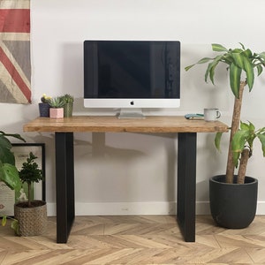 Puede incluir: Un escritorio de madera rústica con patas de metal negro. El escritorio tiene un monitor de computadora, una taza y algunas plantas en macetas en la parte superior. El escritorio está frente a una pared blanca con una bandera de la Unión Jack colgada.
