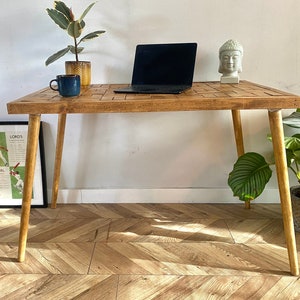 Puede incluir: Una mesa de madera con un acabado natural y una tapa tejida. La mesa tiene cuatro patas y está decorada con una pequeña planta en maceta, una taza azul, un portátil y una estatua de Buda blanca.