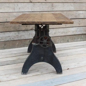 May include: A rustic wooden table top with a black metal base featuring a crank mechanism. The table has a distressed wood finish and a unique industrial design.