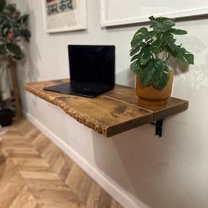 Könnte beinhalten: Ein Wandregal aus Holz mit einem Laptop und einer Topfpflanze darauf. Das Regal hat eine natürliche Holzoptik und eine leicht unebene Kante. Die Pflanze ist eine grüne Monstera in einem braunen Topf.