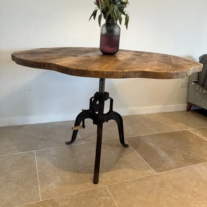 Könnte beinhalten: Ein rustikaler Holztisch mit einer einzigartigen, unregelmäßigen Form und einem schwarzen Metallgestell mit einer Handkurbel. Der Tisch steht auf einem hellbraunen Fliesenboden und hat eine Blumenvase darauf.